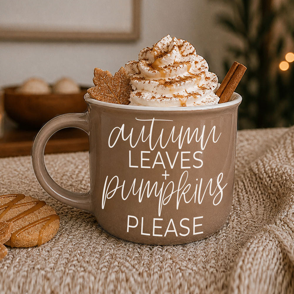 Brown mug with 'Autumn leaves + pumpkins please' text, topped with whipped cream and a cinnamon stick, on a textured surface.