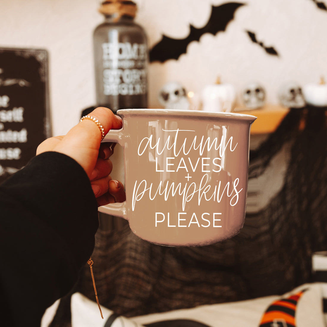Person holding a mug with 'autumn leaves pumpkins please' text in a Halloween-themed setting.
