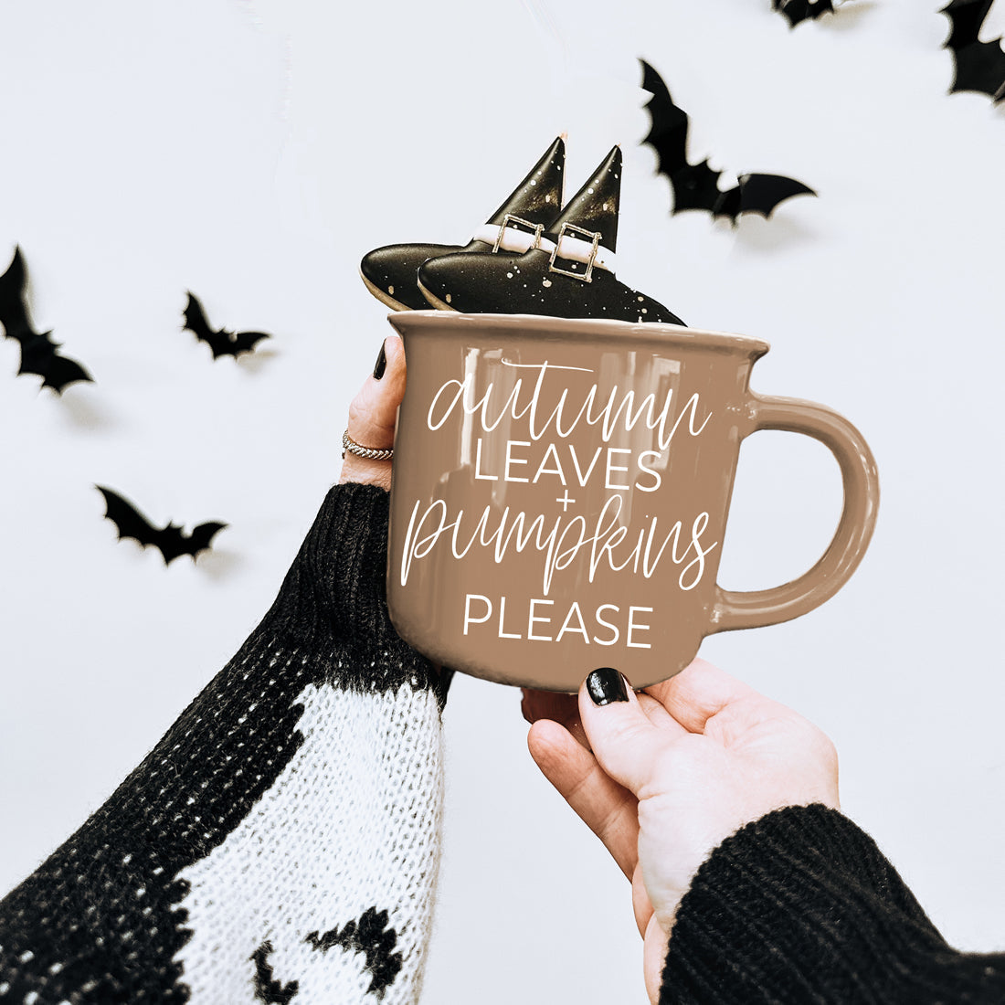 Person holding a mug with 'Autumn leaves + pumpkins please' text against a Halloween-themed background with bats.