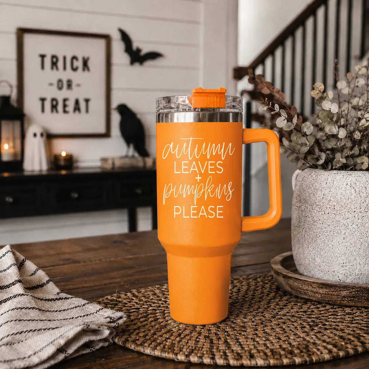 Orange tumbler with text on a table in a cozy room with Halloween decorations.
