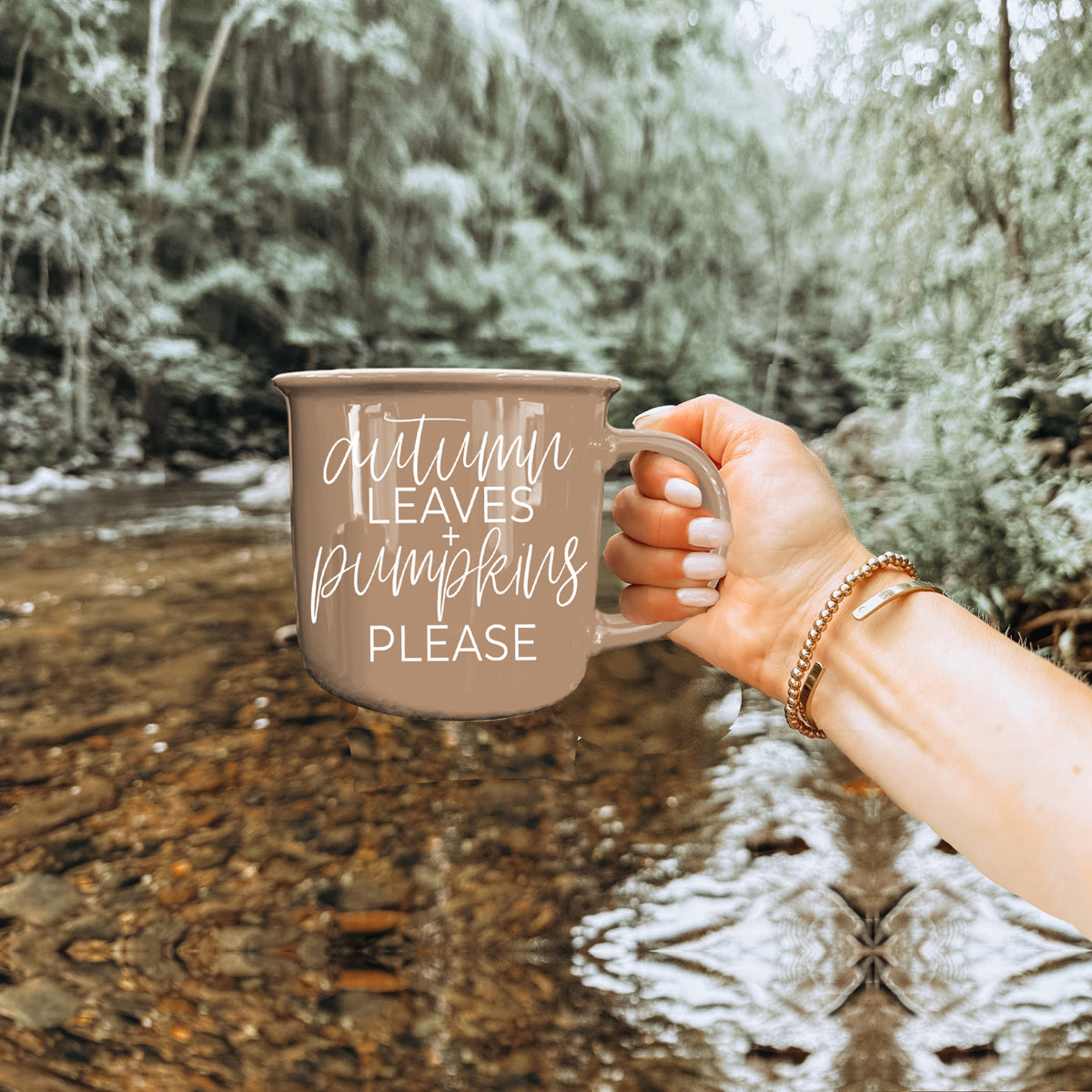 Hand holding a mug with 'Autumn leaves pumpkins please' text in a forest setting