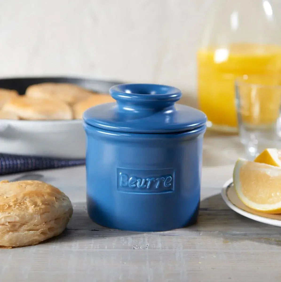 Blue ceramic butter crock labeled 'Beurre' on rustic table with biscuits and orange juice, farmhouse kitchen decor