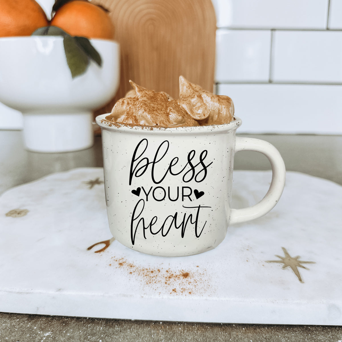 Mug with 'bless your heart' text, topped with whipped cream and cinnamon, on a kitchen counter.