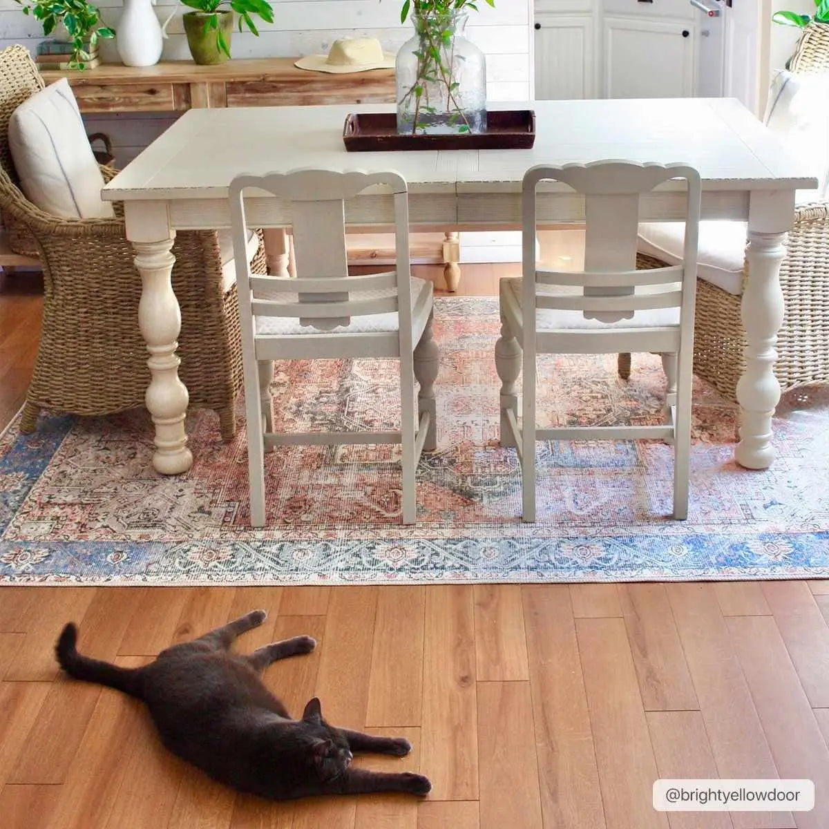 Cozy farmhouse dining table with white chairs, woven accents, and a gray cat lounging on wood floor