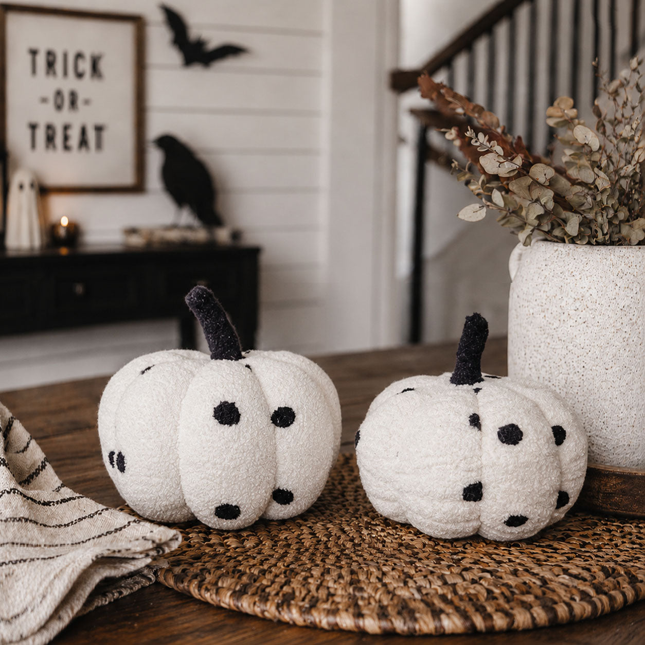 White pumpkins with black spots on a woven mat, Halloween decorations in the background.