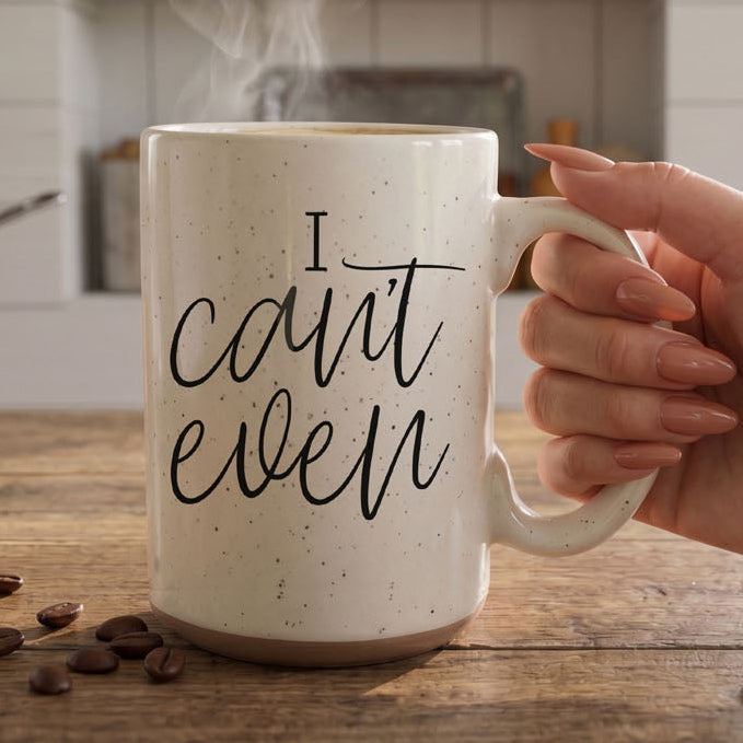 Mug with 'I can't even' text held by a hand on a wooden surface with coffee beans.