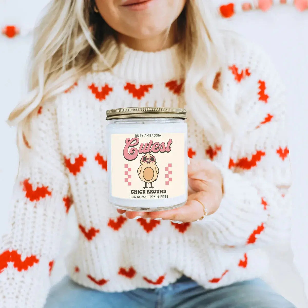 Person holding a candle labeled 'Cutest Chick Around' in a cozy setting