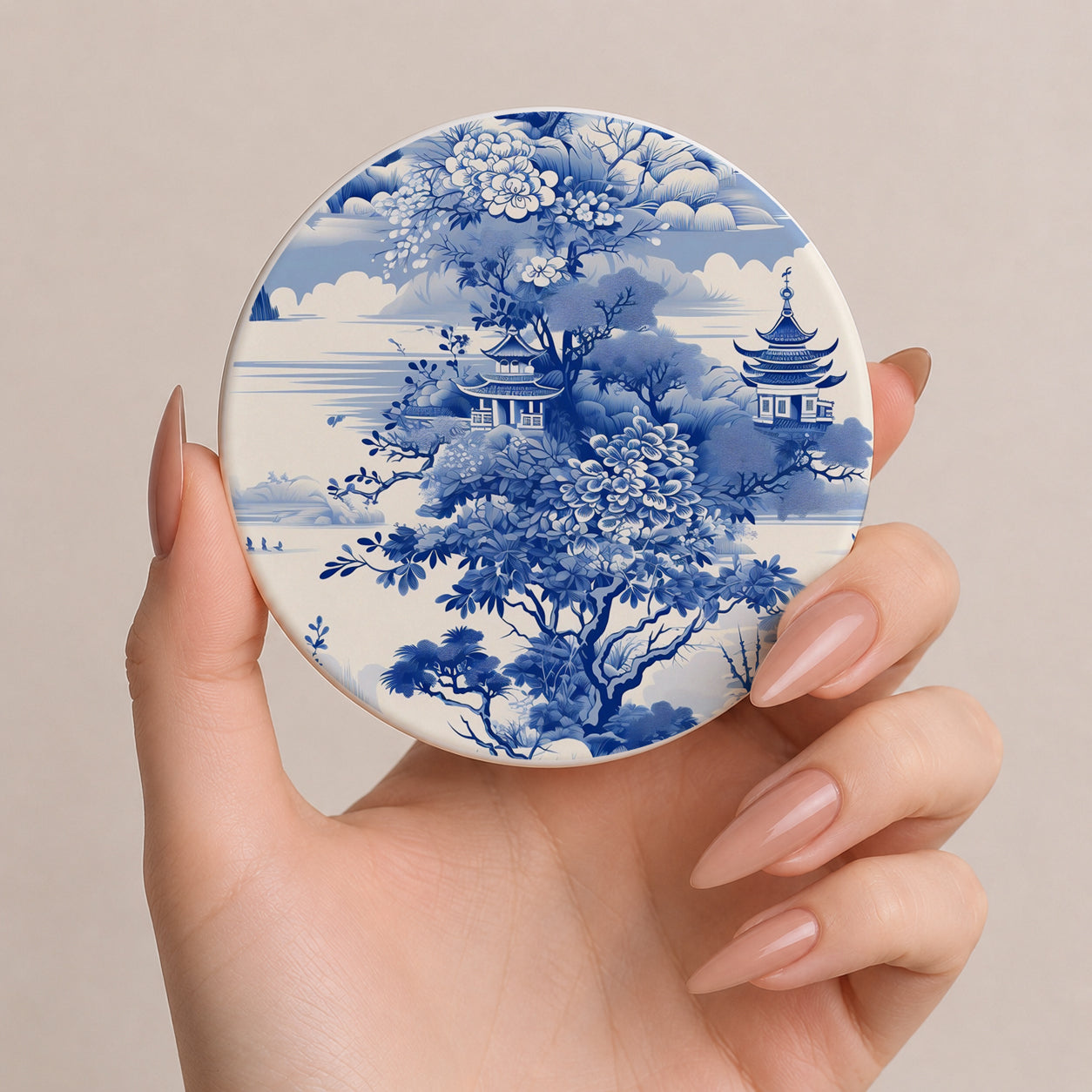 Hand holding a round compact mirror with blue floral and architectural design on a beige background