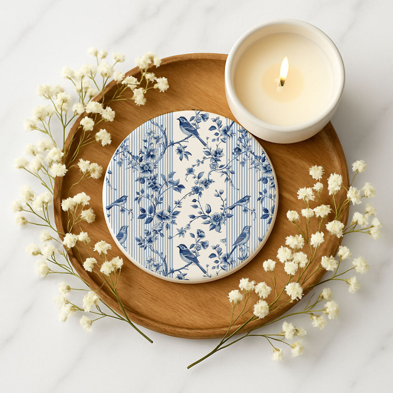 Decorative coaster with blue floral and bird pattern on a wooden tray with a candle and flowers.