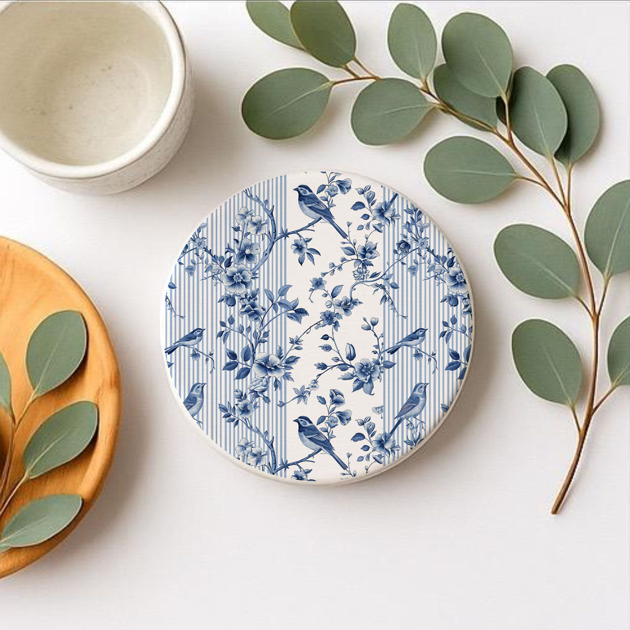 Round ceramic coaster with blue floral and bird pattern on a white surface with green leaves.