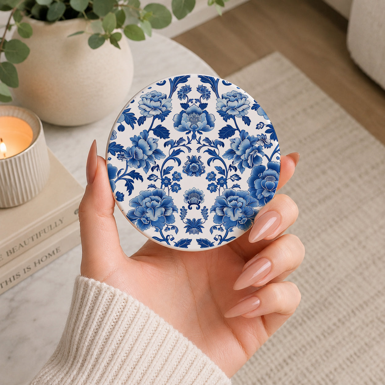 Hand holding a round blue floral patterned coaster with a cozy indoor setting in the background.
