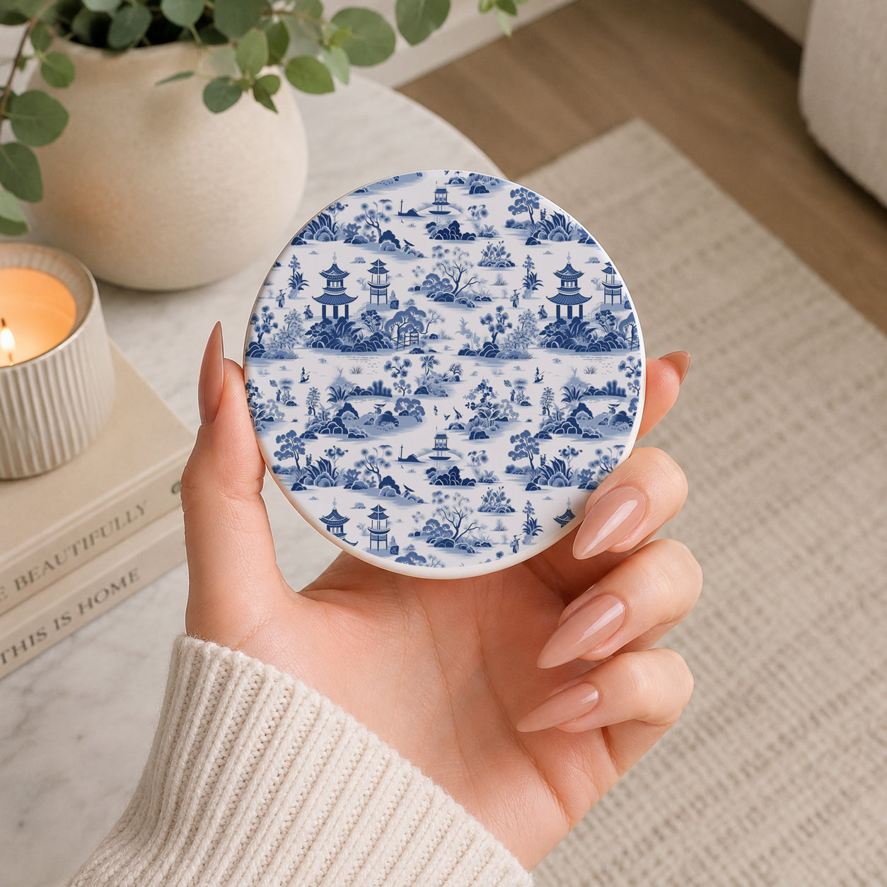 Hand holding a round ceramic coaster with blue and white pattern, blurred indoor setting with books and plant.