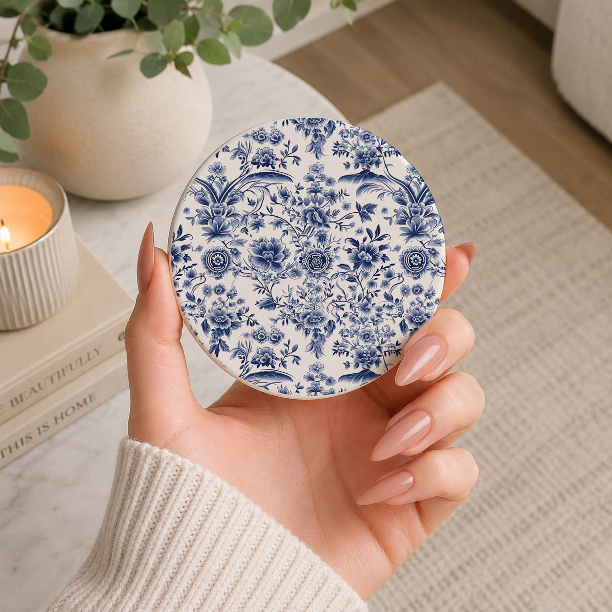 Hand holding a blue floral patterned coaster with a cozy indoor setting in the background.
