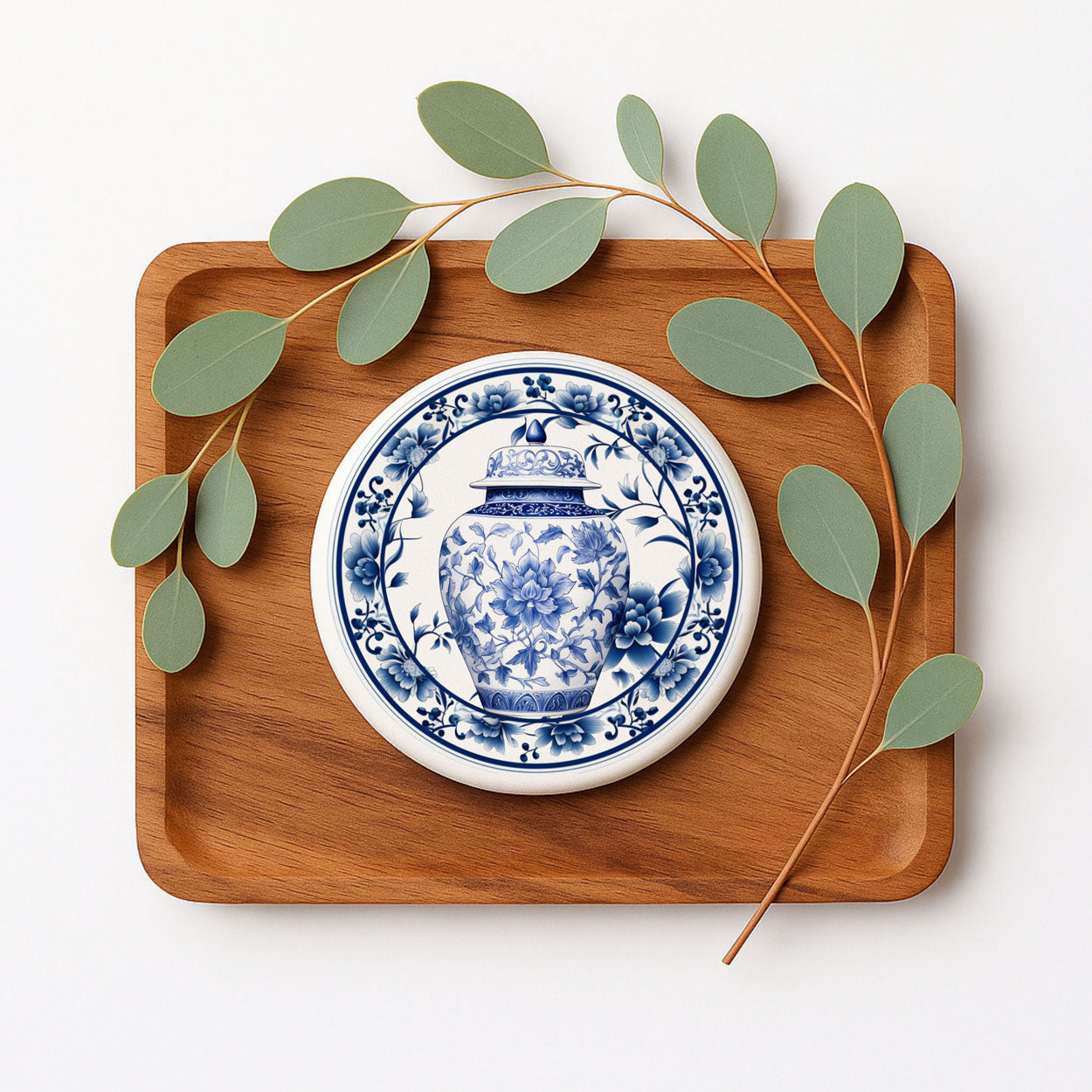 Wooden tray with a blue and white ceramic coaster featuring a floral design, surrounded by green leaves on a white background.