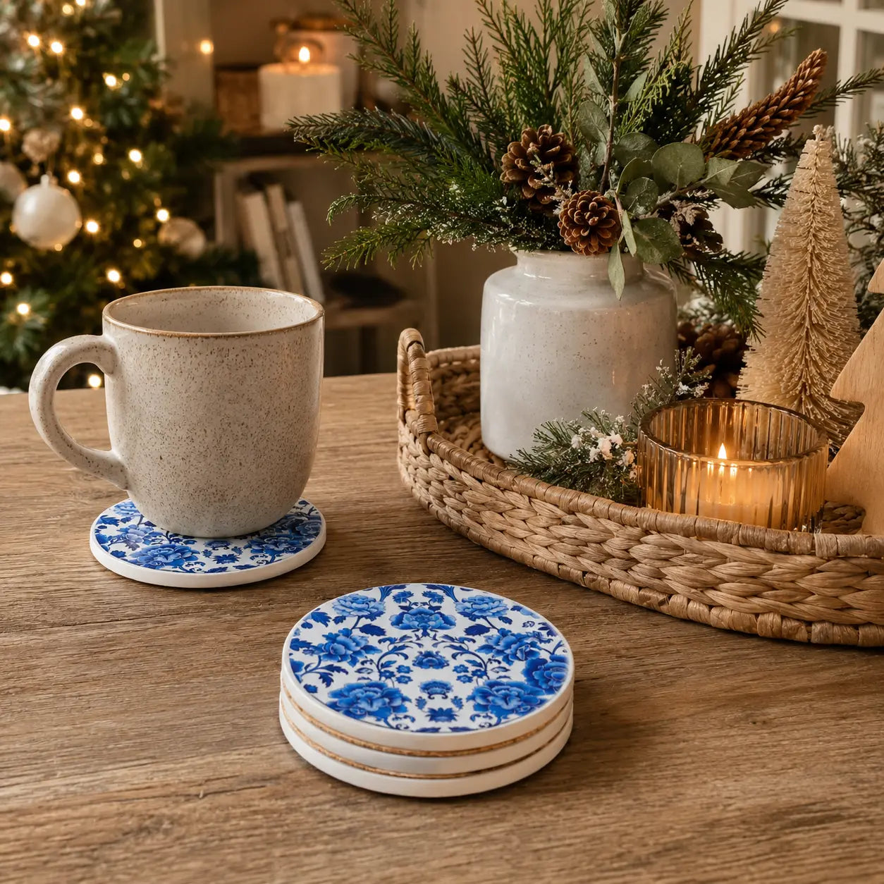 Blue and white floral patterned ceramic coasters on a wooden table, styled with a speckled beige mug and surrounded by holiday decor in a cozy indoor setting.