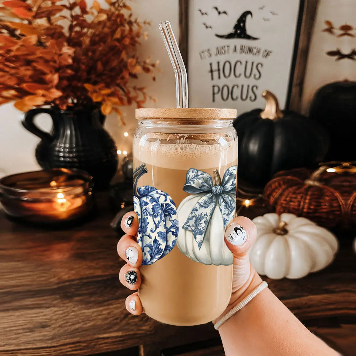 Hand holding a mason jar with a pumpkin spice drink, decorated with blue and white pumpkins and bows, against a Halloween-themed background.