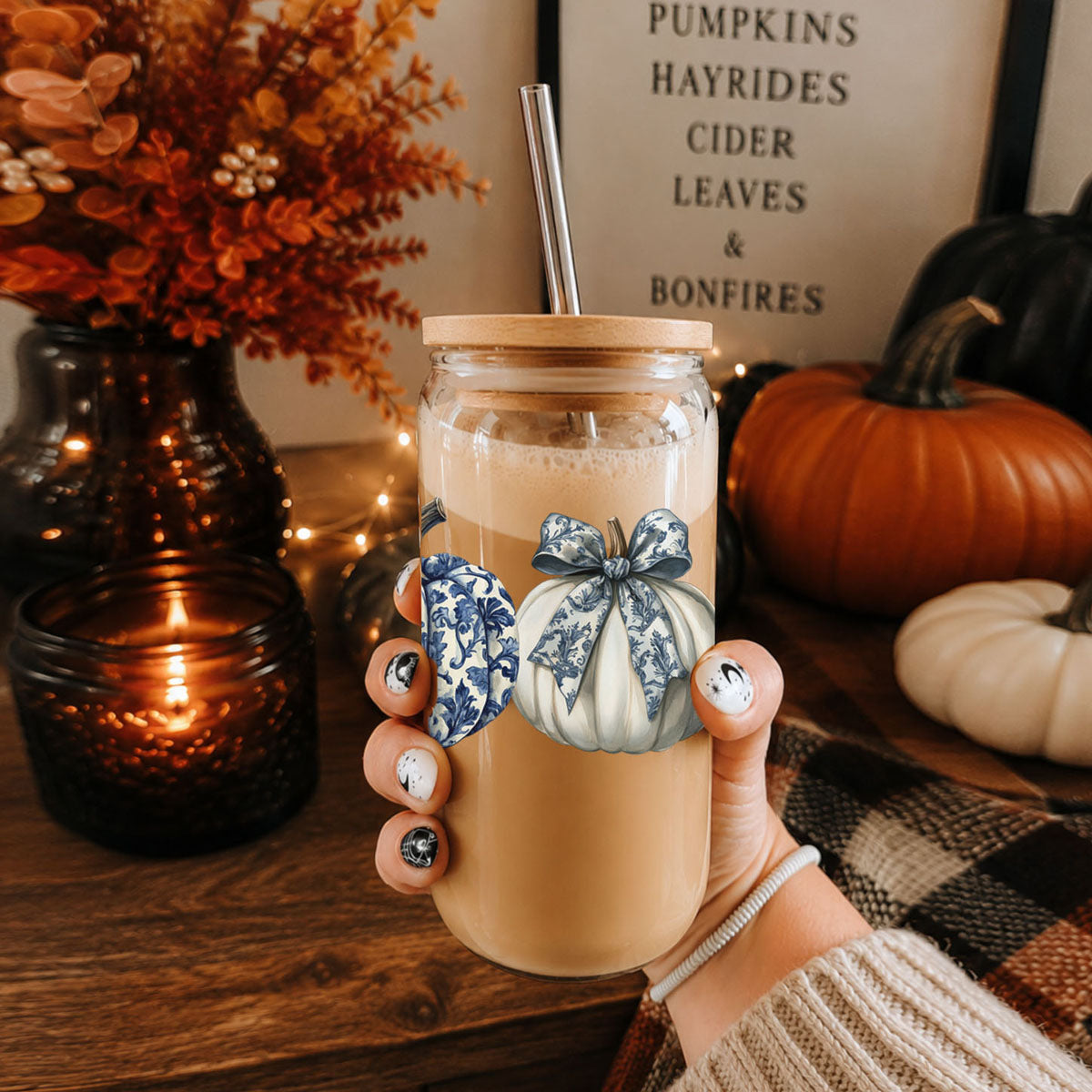 Hand holding a mason jar with a pumpkin spice latte, surrounded by autumn decorations.