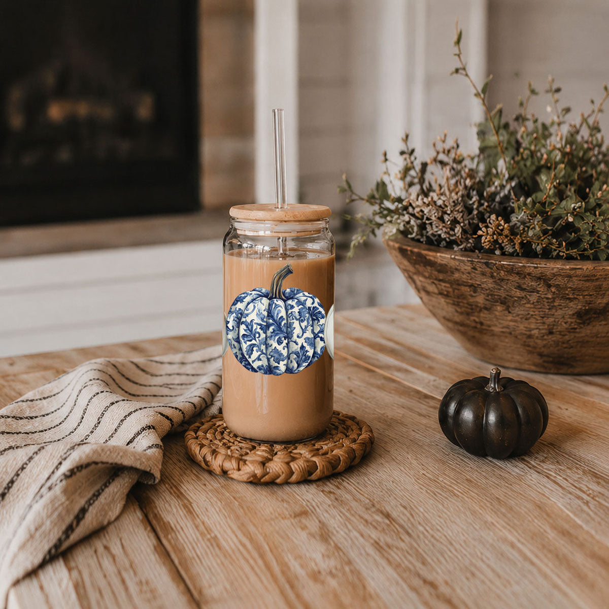Glass tumbler with a pumpkin design on a wooden surface with a bowl of flowers and a small pumpkin.