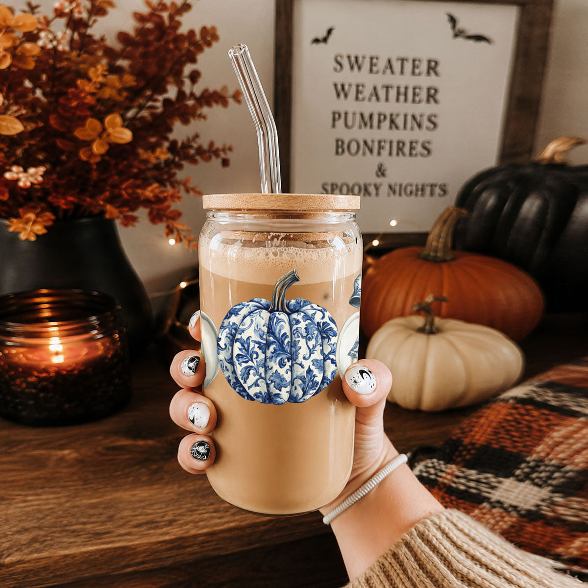 Hand holding a glass of iced coffee with a decorative pumpkin, autumn decorations in the background.