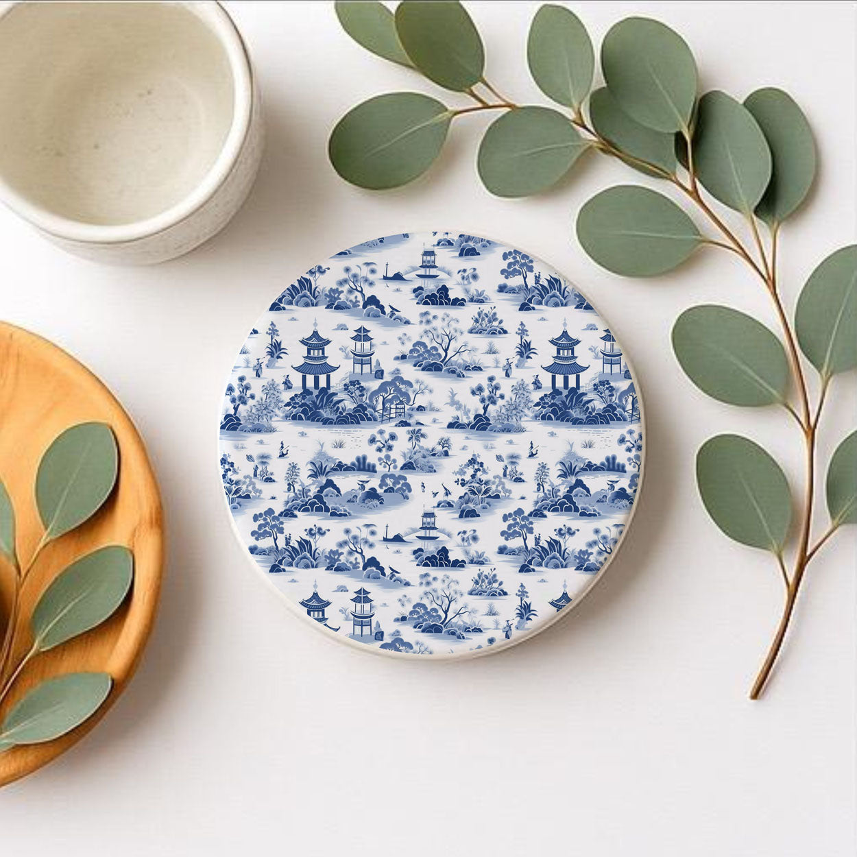 Round ceramic coaster with blue and white pattern on a white surface with a wooden plate and green leaves.