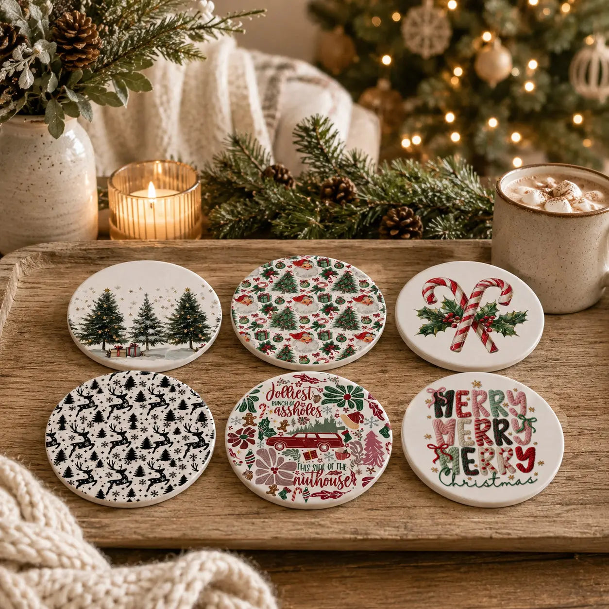 Six round ceramic coasters with festive Christmas designs are arranged on a wooden tray in a cozy holiday setting with a candle, mug, pine branches, and blurred Christmas tree lights in the background.