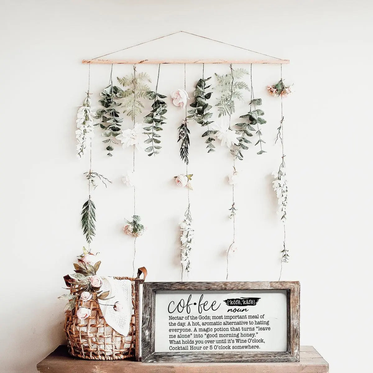 Decorative wall hanging with plants and a framed definition of 'coffee' on a white wall.