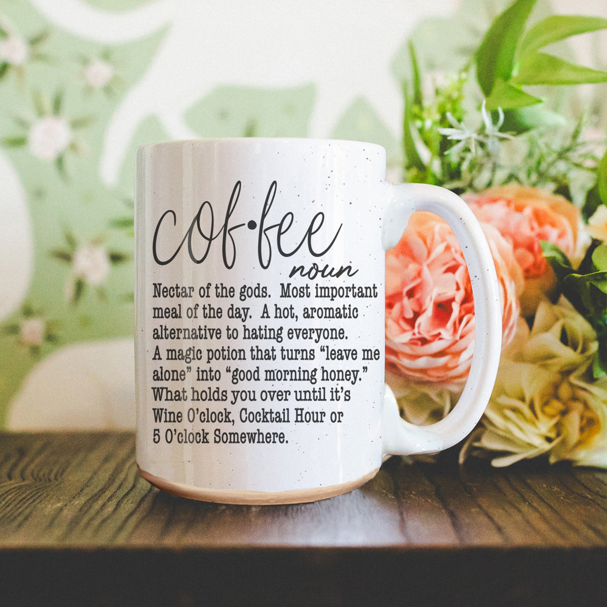 White mug with coffee definition text on a wooden surface with flowers in the background