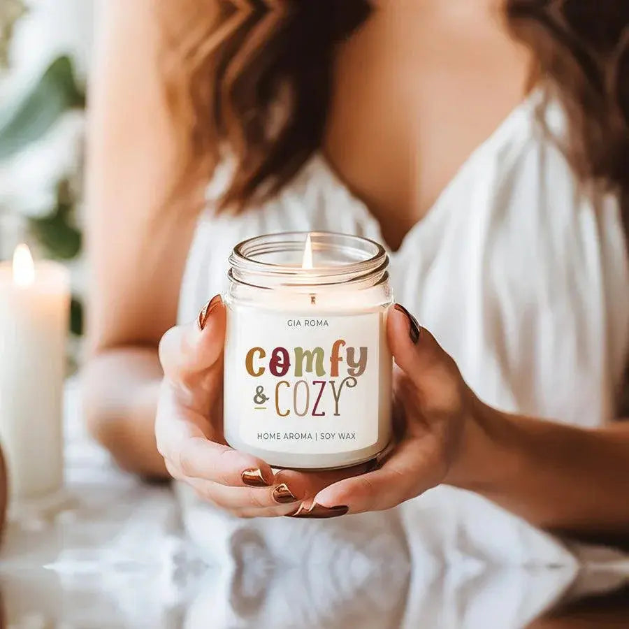 Person holding a 'comfy & cozy' candle jar with another candle in the background.