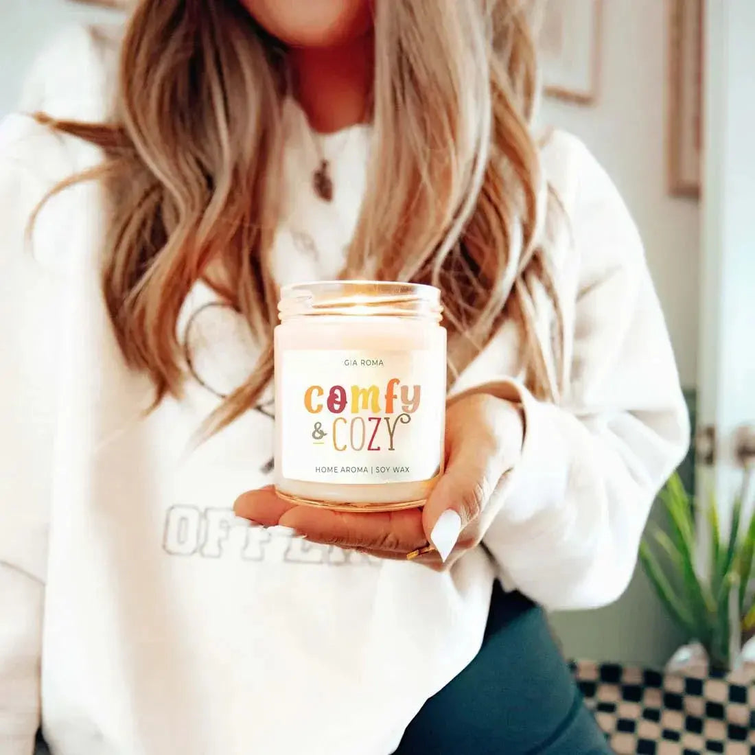 Person holding a 'comfy & cozy' candle in a home setting