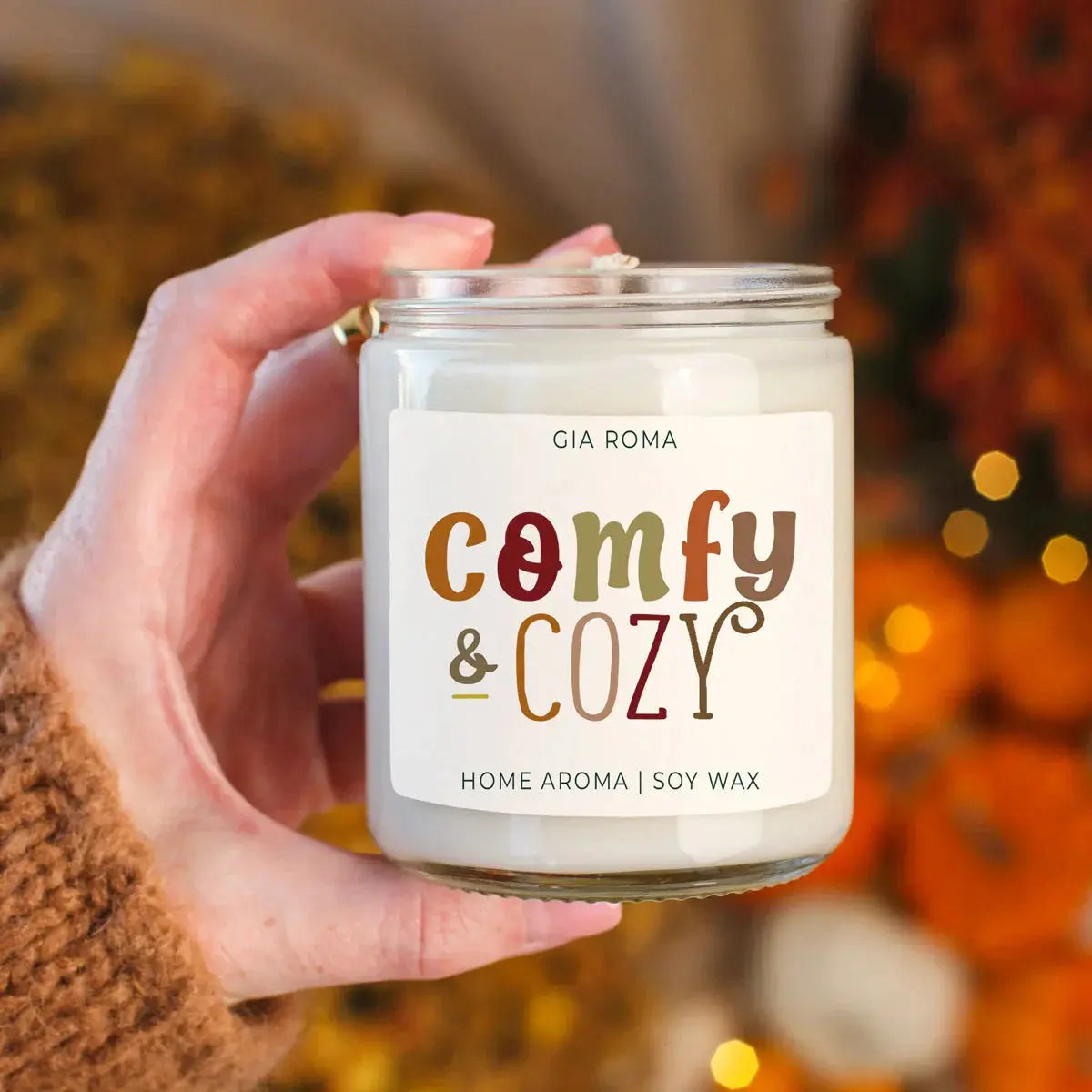Hand holding a candle labeled 'Comfy & Cozy' with a blurred autumn background