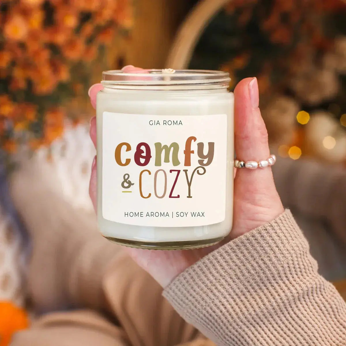 Hand holding a 'Comfy & Cozy' candle with blurred autumn background