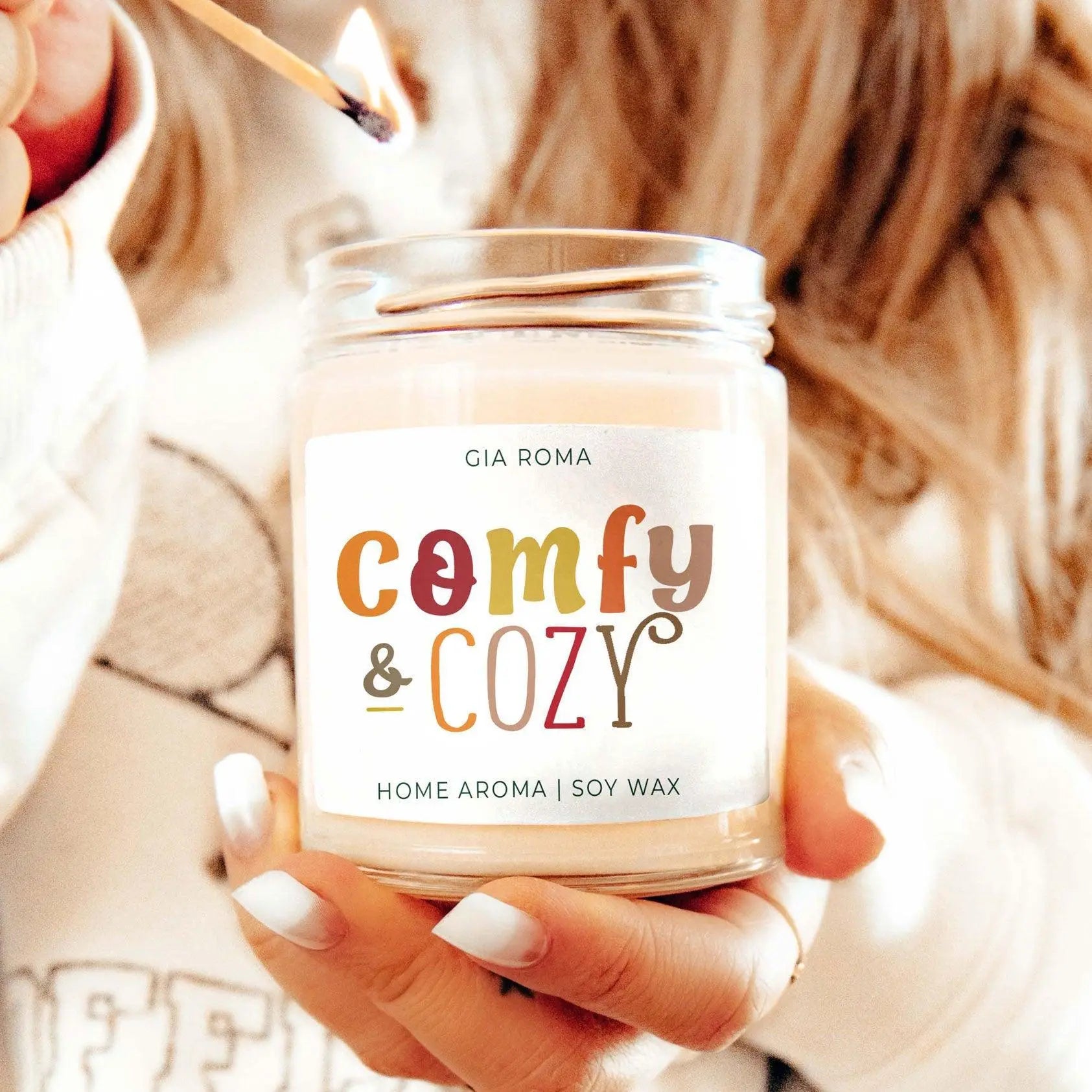 Candle labeled 'comfy & cozy' by Gia Roma held by a person with a match.