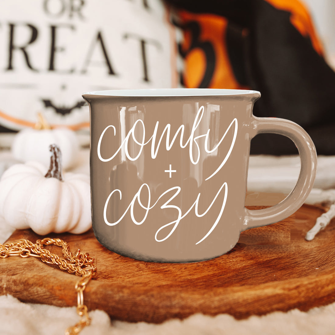 Gray mug with 'comfy + cozy' text on a wooden surface with pumpkins and a blurred background.
