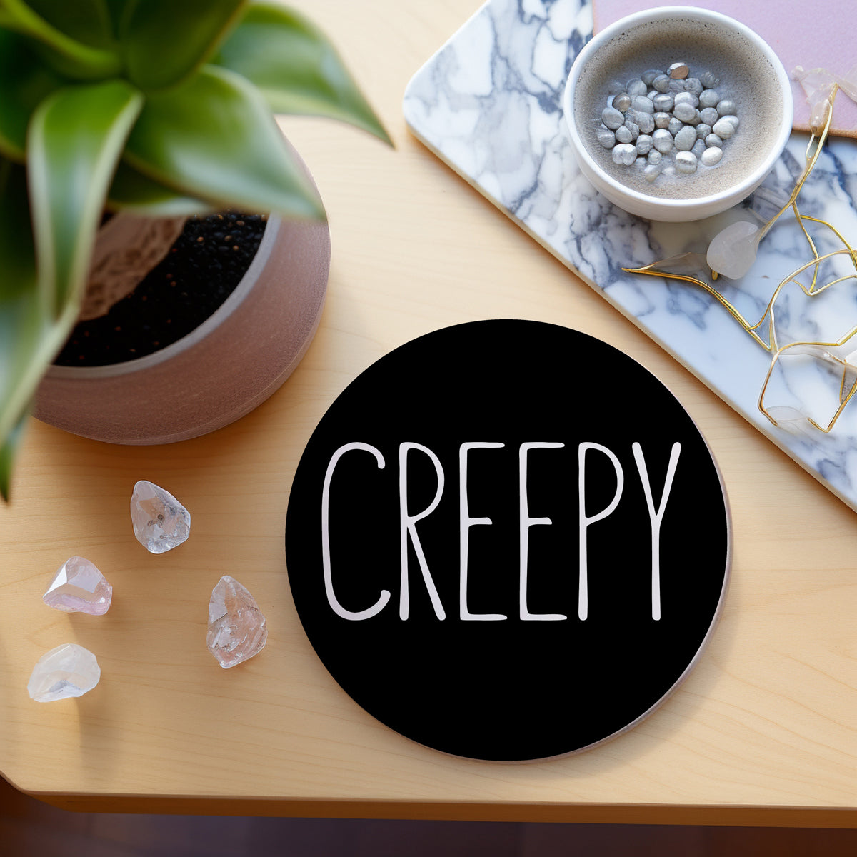 Round black coaster with 'CREEPY' text on a wooden surface with decorative items.