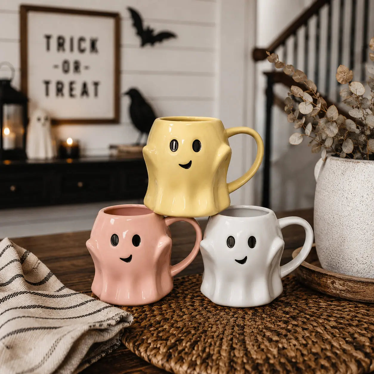 Three ghost-shaped mugs in yellow, pink, and white on a table with Halloween decorations in the background.