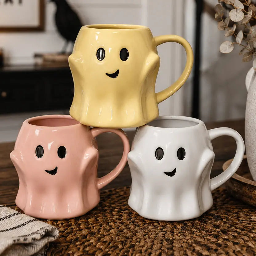 Three ghost-shaped mugs in yellow, pink, and white on a textured surface.