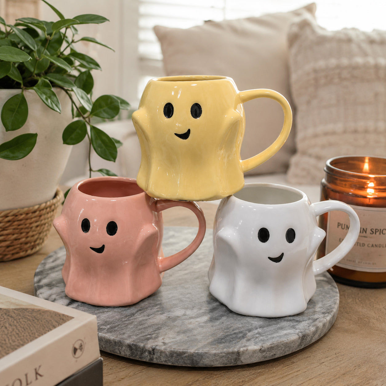 Three colorful ghost-shaped mugs on a marble coaster with a candle and plant in the background.
