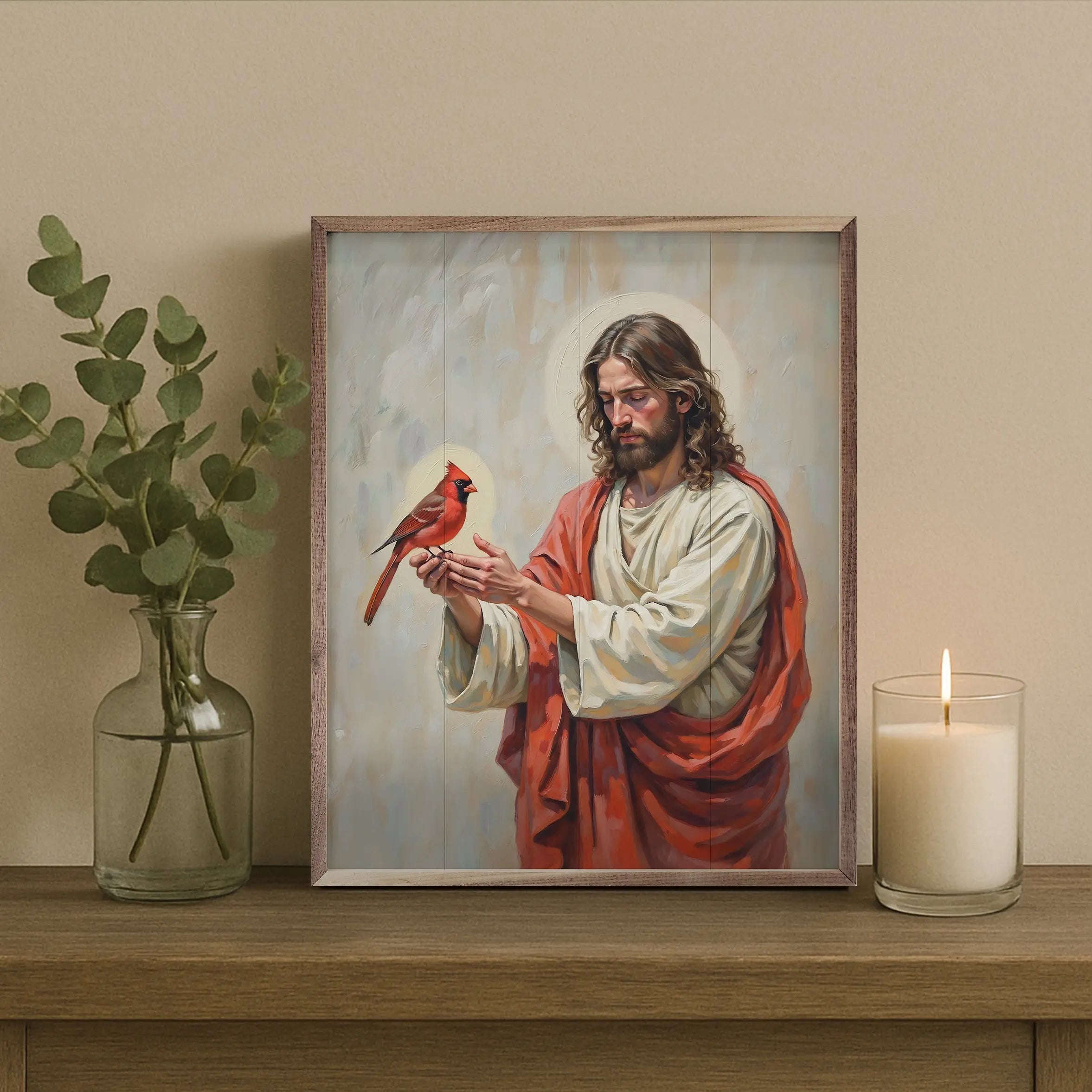 Framed artwork of a religious figure holding a cardinal bird on a wooden surface with a candle and plant.