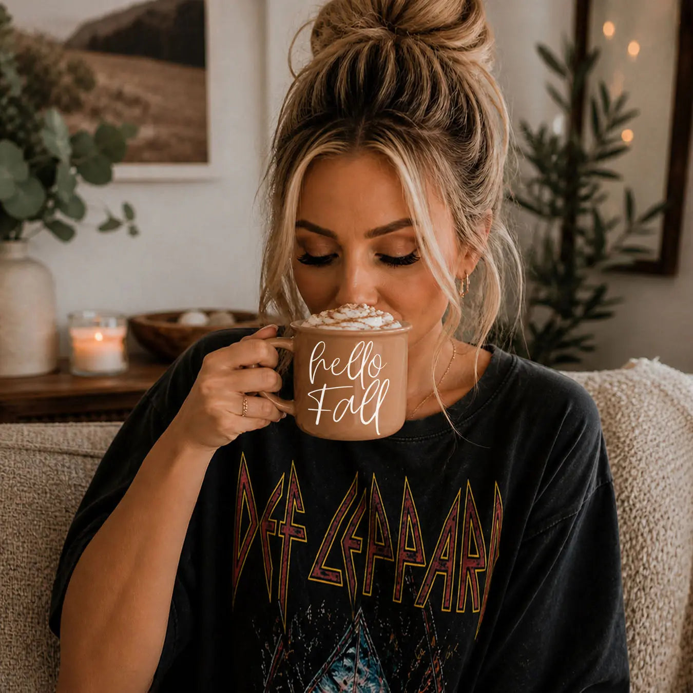 Woman holding a mug with 'Sweater Weather' text, wearing a Def Leppard t-shirt, in a cozy indoor setting.