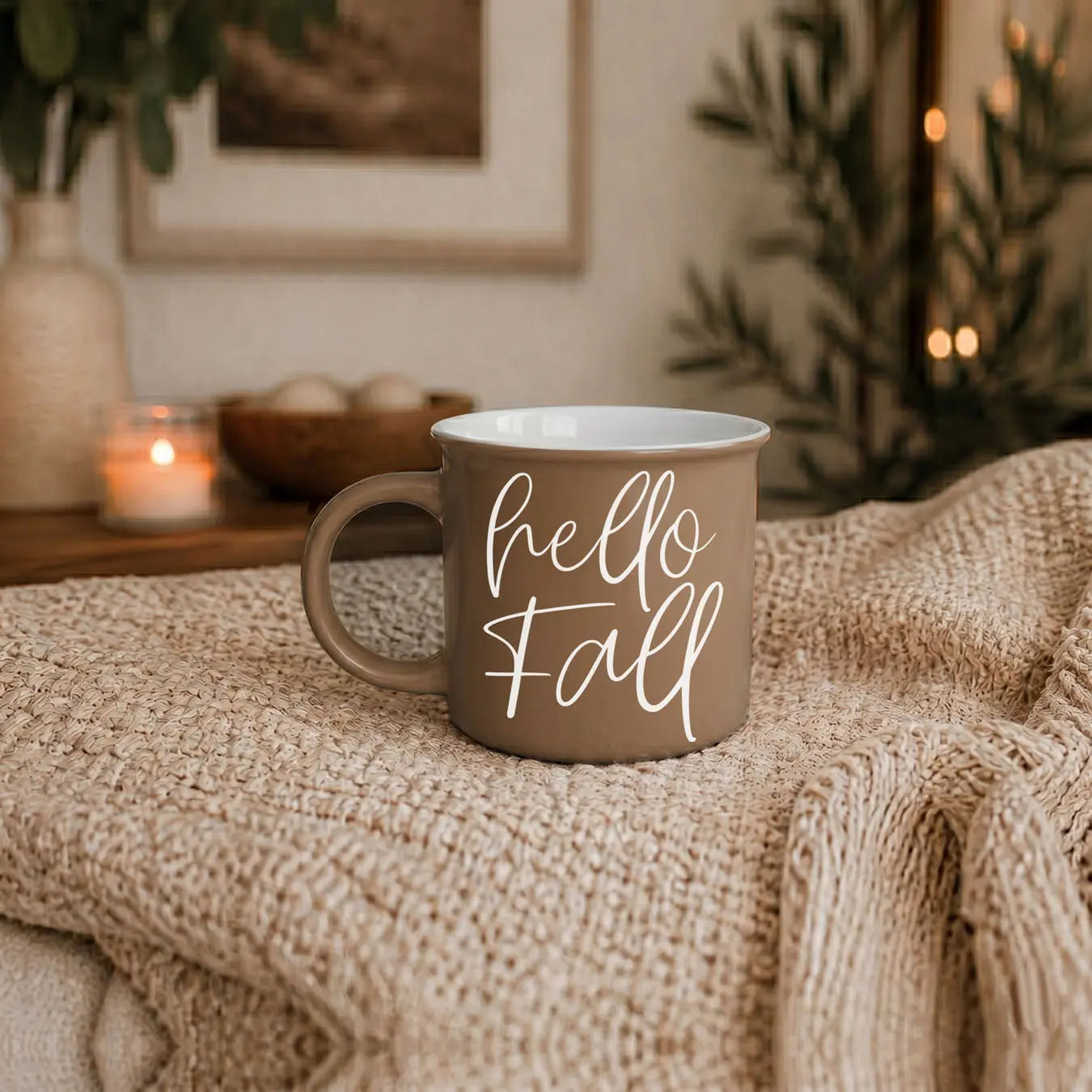 Mug with 'hello fall' text on a textured surface with candles and plants in the background
