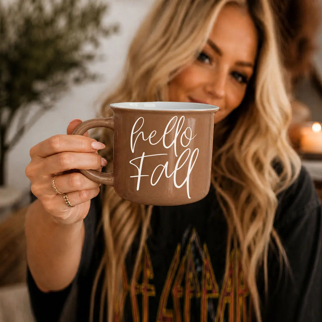 A woman holds up a brown ceramic mug with white text that reads 'Fall MY 2ND FAVORITE f-word' against a warm, softly blurred indoor background.