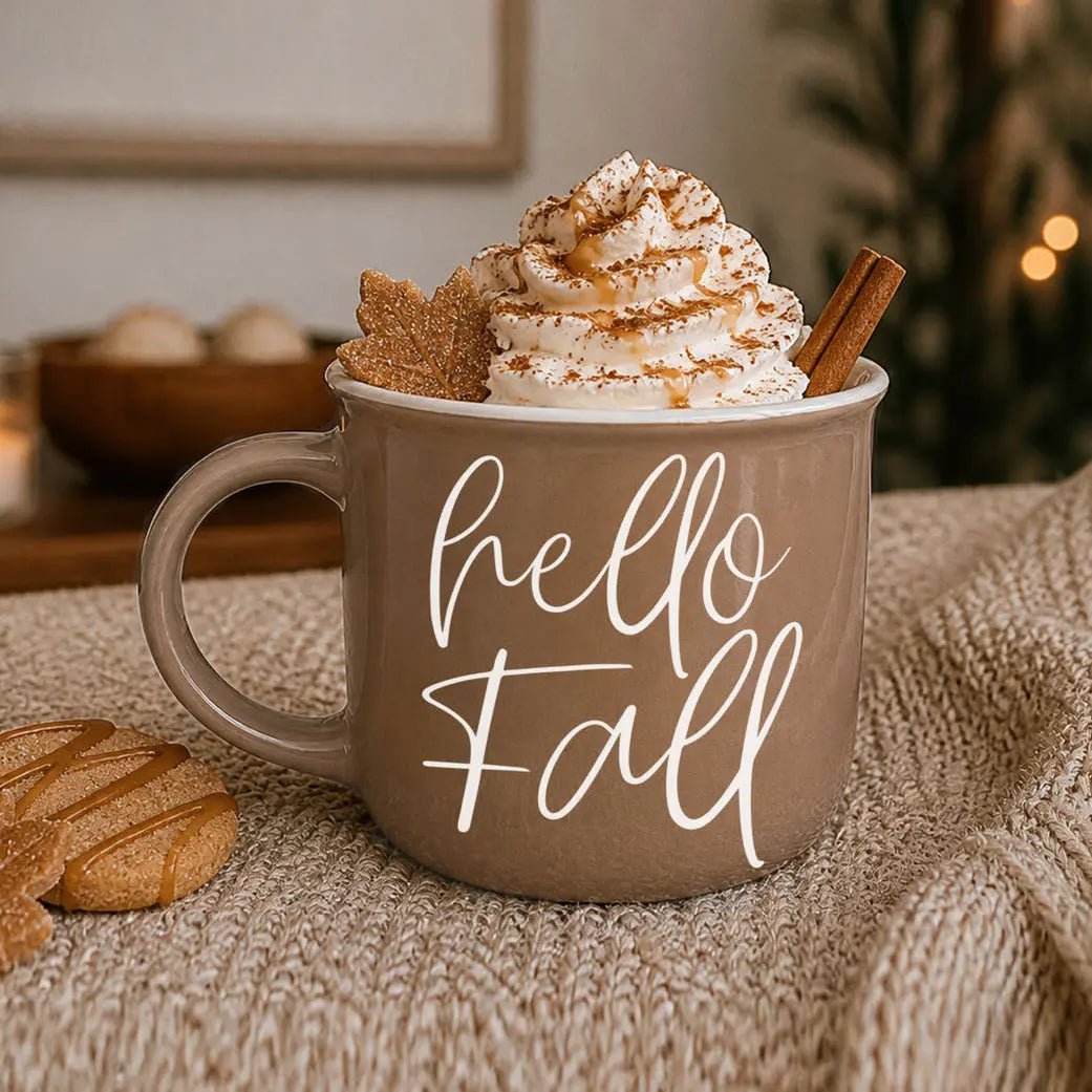 Brown mug with 'hello Fall' text, topped with whipped cream and a cinnamon stick, on a textured surface.