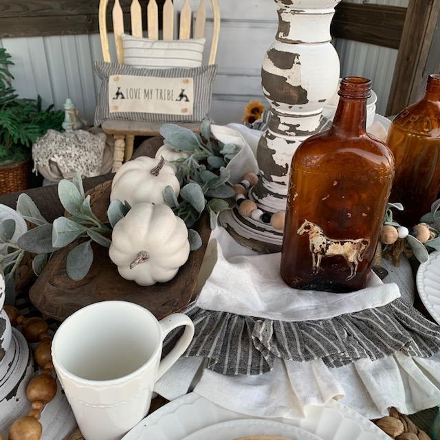 A styled rustic farmhouse table setting featuring an amber glass bottle with a cow motif, white ceramic mugs and plates, sage greenery, small white pumpkins, and a distressed white and brown candle holder.