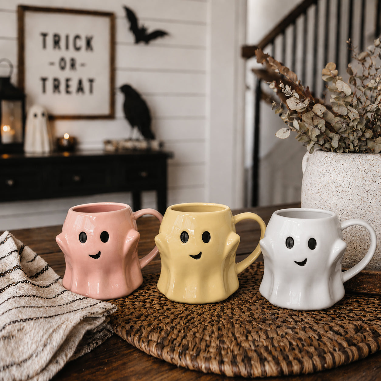 Three ghost-shaped mugs in pink, yellow, and white on a woven mat with Halloween decorations in the background.