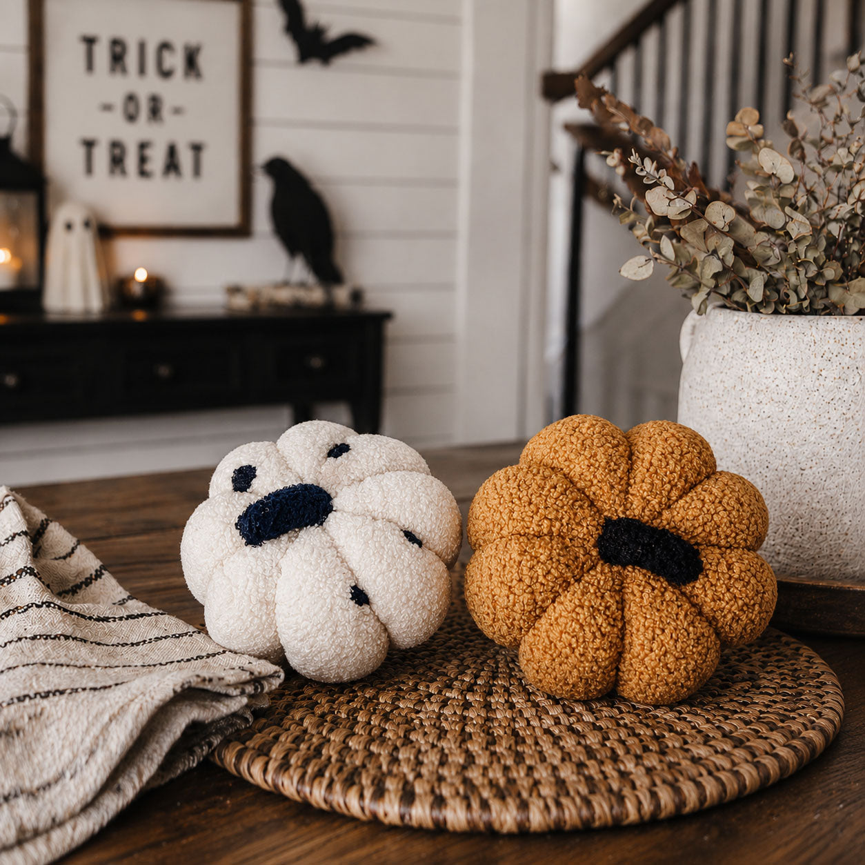 Decorative pumpkins on a woven mat with Halloween-themed decor in the background.
