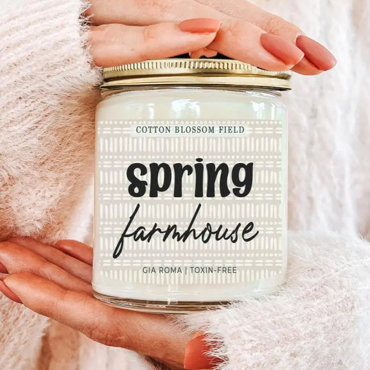 Candle labeled 'Spring Farmhouse' held by a hand on a soft pink background