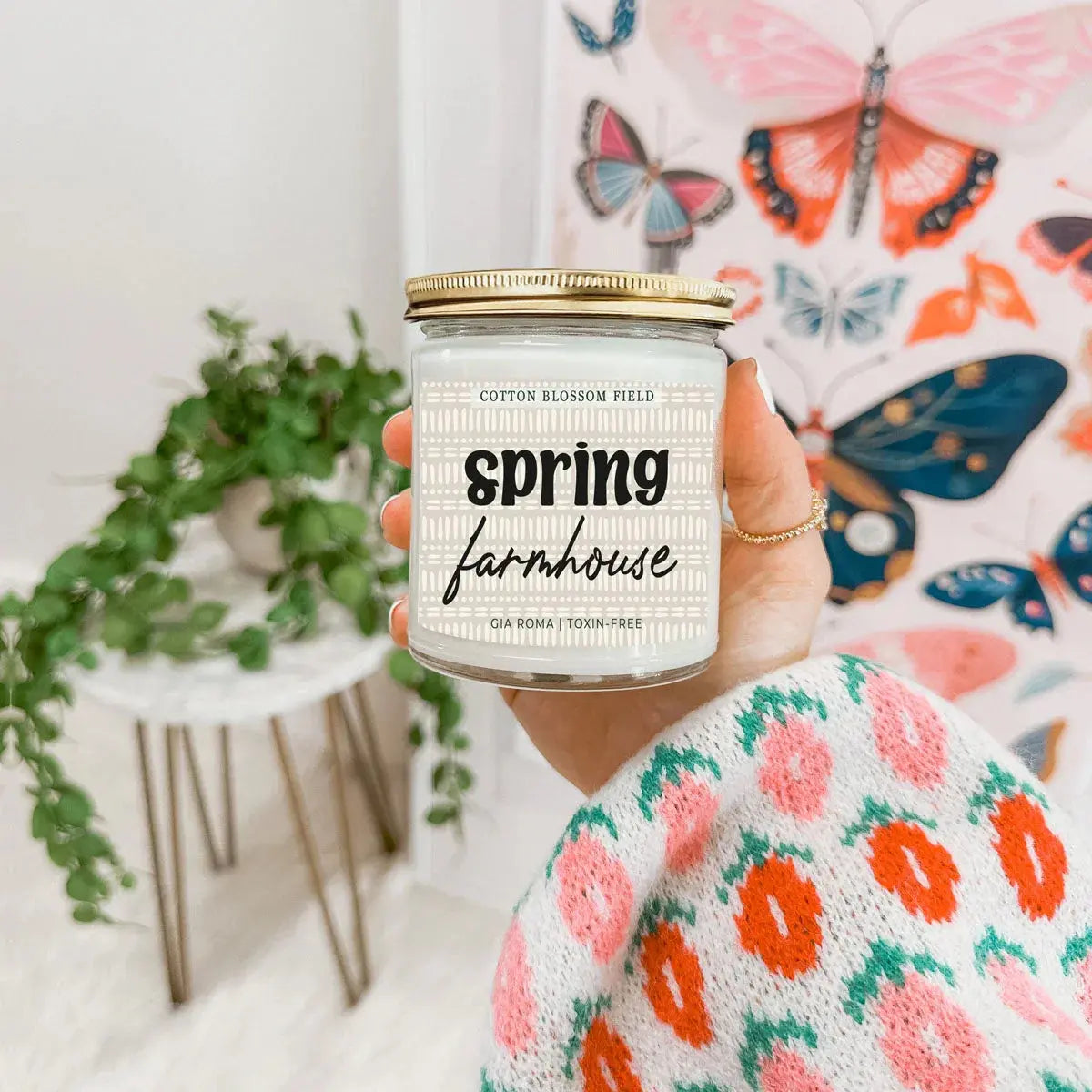 Hand holding a 'Spring Farmhouse' candle with a colorful butterfly-themed background