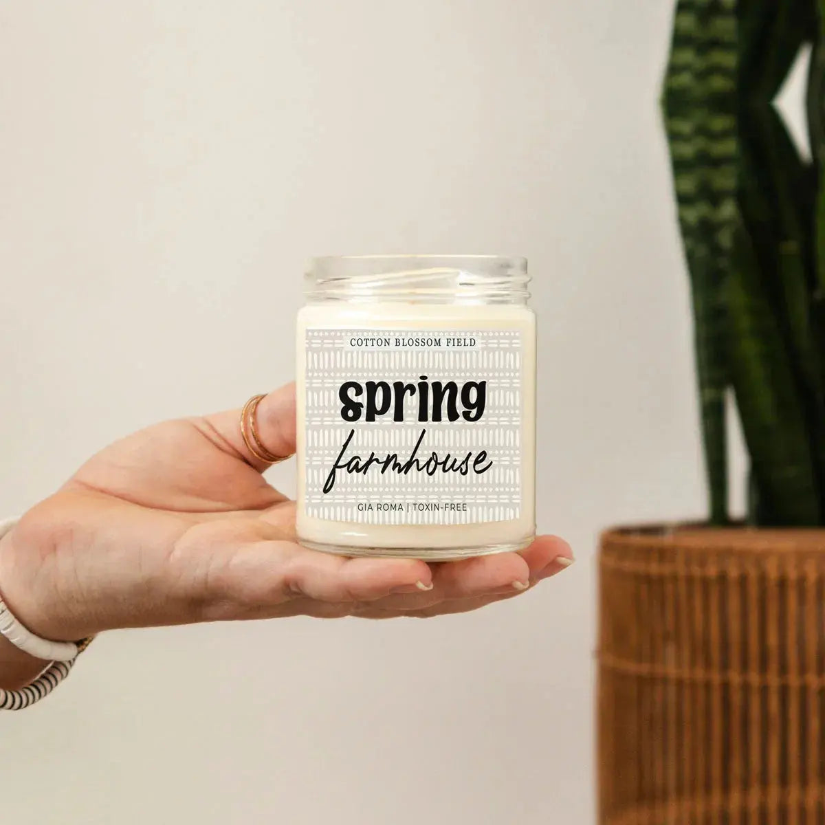 Hand holding a candle labeled 'spring farmhouse' with a plant in the background