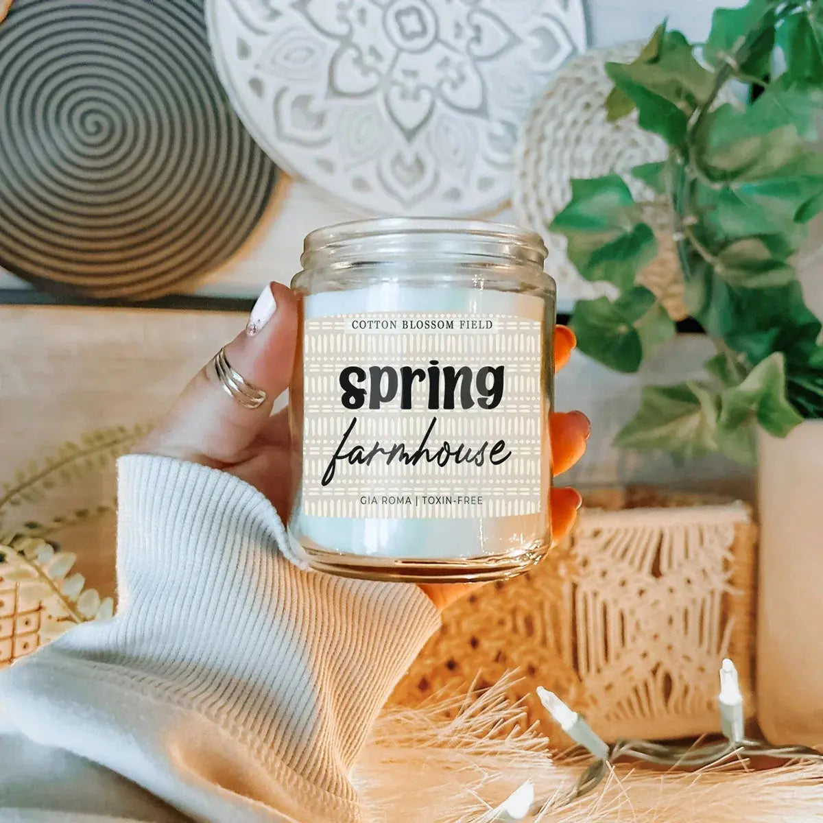 Hand holding a candle labeled 'Spring Farmhouse' with decorative background