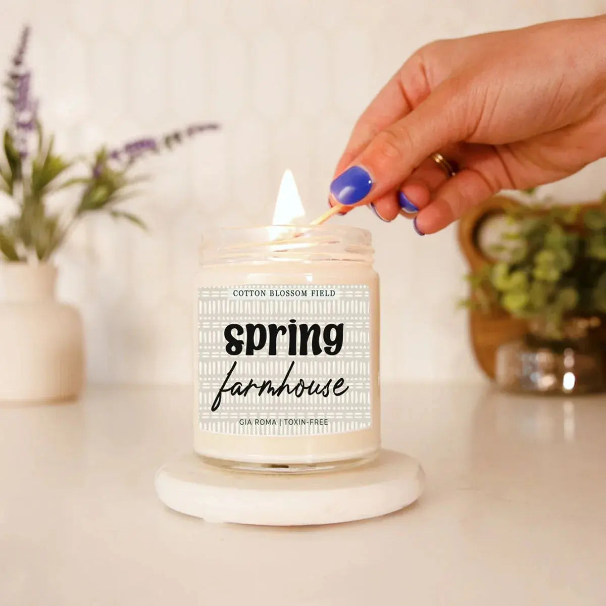 Candle labeled 'Spring Farmhouse' being lit with a match, surrounded by decorative plants.